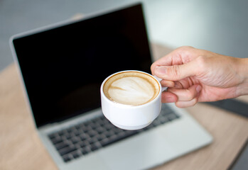 A cup of latte art coffee in hand. Laptop computer with blank screen are on the background.