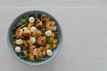 Top view of salad with mango, mozzarella, shrimps and mixed microgreens in blue bowl on white oak table