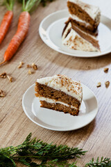 piece of carrot cake on a white plate on a wooden table. Two carrots and walnuts for decoration.