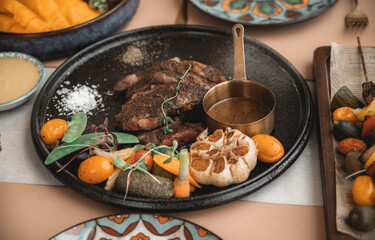 Cooked medium raw beef steak with blood sauce salt and pepper on black rough clay plates served with roasted garlic, peas, tomatoes and carrots. Flat lay between other dished and clutter ready to eat