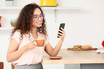 Fototapeta na wymiar Beautiful young woman with cup of coffee using mobile phone in kitchen