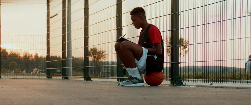 Black African American teenager boy writing lyrics for his song on a basketball court. Aspiring rap artist dreaming of becoming future star. Shot with 2x anamorphic lens