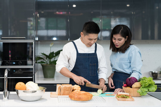 A happy man and young woman cut vegetables to cook healthy sandwiches on the kitchen island. Asian couple uses their free time to cooking food together on weekends in the kitchen. Culinary lifestyle.