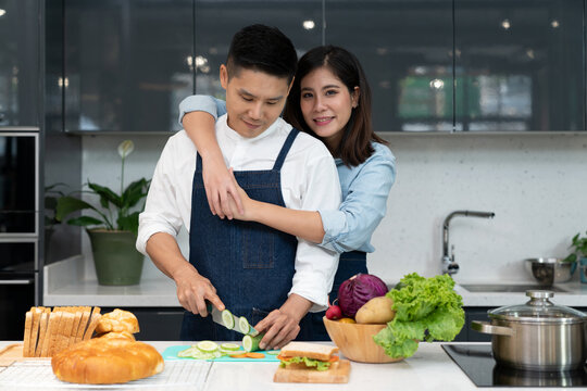 A happy man and young woman cut vegetables to cook healthy sandwiches on the kitchen island. Asian couple uses their free time to cooking food together on weekends in the kitchen. Culinary lifestyle.