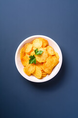 Fried corrugated golden potato chips with parsley leaf in wooden bowl on blue background, top view