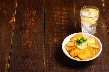 Fresh ice cold water drink with lemon near to fried potato chips with parsley leaf in wooden bowl on wood backdrop, angle view copy space