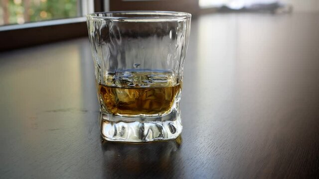 Close-up of mixing whiskey and ice cubes in a glass on a wooden table. A glass of ice and scotch whiskey. A drop of alcohol runs down the side of the glass.