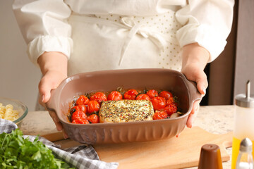 Woman holding dish with baked tomatoes and feta cheese in kitchen - obrazy, fototapety, plakaty