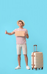 Young man with cardboard and suitcase hitchhiking on color background