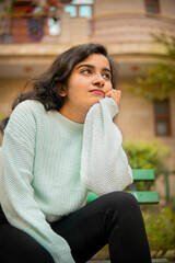 Fototapeta premium Outdoor low angle image of an Asian, Indian beautiful happy young woman sitting on park bench and contemplating while looking away with toothy smile and enjoying fresh air at day time.