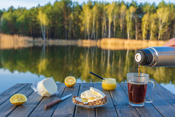 Pouring hot tea into a glass mug from a thermos in the morning next to the lake and forest. Breakfast on a wooden table