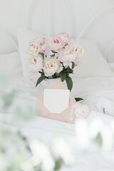 Bouquet of pink peonies and greeting envelope on white bed linen. Modern interior in the bedroom. Wedding and festive style.