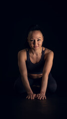 Fototapeta na wymiar Studio photo, with black background, of an Asian woman, fitness instructor, in sports clothes, sitting on her knees, looking at the camera smiling, with a confident expression.