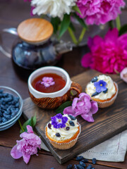 Obraz na płótnie Canvas Cupcake with cream decorated with violet flowers lying next to a mug of tea among the flowers of peonies and scattered honeysuckle berries
