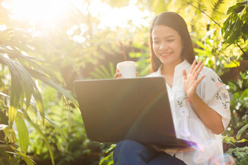 Beautiful young woman enjoying morning coffee and making a video call to friends in the garden at home. Asian female say hi and waving to their family while using laptop in a back garden.

P