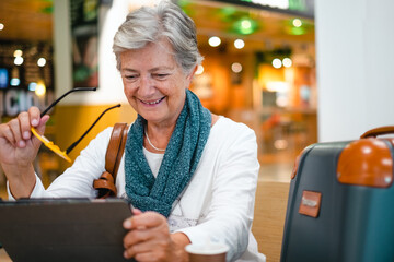 Senior woman sitting at the airport cafe using a digital tablet while waiting for boarding. Happy mature traveler drinks coffee