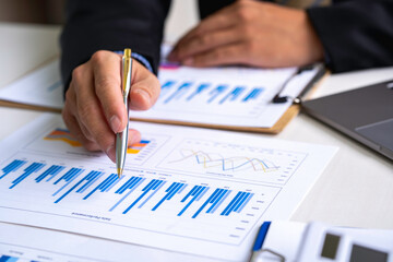 Businessman holding pen and pointing paper chart summary analyzing annual business report with using laptop at room office desk.