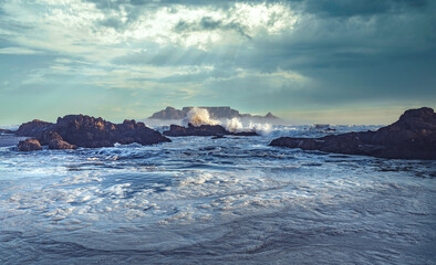 Waves crash on the rocks of Big Bay beach with iconic Table Mountain in the background, Cape Town, South Africa.