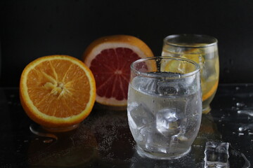 Ice cube flies falls into the takan with water soda next to lie orange grapefruit and ice on a black background. Refreshing drinks. Lemonade, fruit water