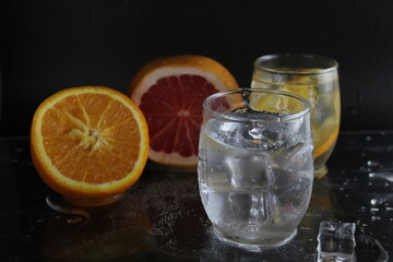 Ice cube flies falls into the takan with water soda next to lie orange grapefruit and ice on a black background. Refreshing drinks. Lemonade, fruit water