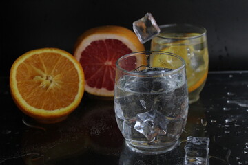 Ice cube flies falls into the takan with water soda next to lie orange grapefruit and ice on a black background. Refreshing drinks. Lemonade, fruit water