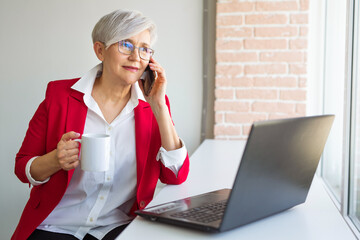 adult elderly woman in a red jacket in an office with a laptop calls on the phone 