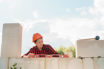 young attractive female builder in a protective helmet, lays aerated concrete blocks on a construction site. Checks the zero level of the walls, using the building level. Copy space