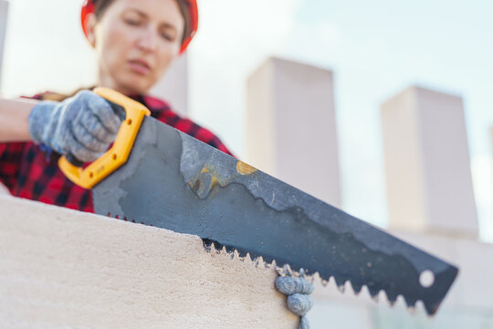 young female construction worker in a protective helmet, sawing aerated concrete blocks with a saw. The process of working as a bricklayer's helper. Copy space