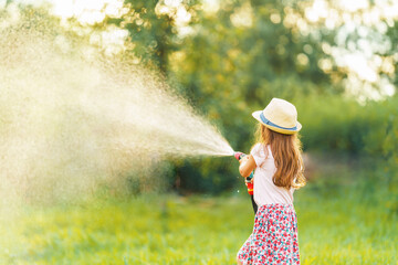 happy little girl is watering plants and playing with garden hose with sprinkler on sunny day in backyard, enjoying splashing water in heat. Summer entertainment and outdoor recreation for children.