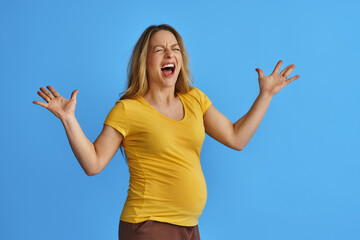 young pregnant woman expecting baby on an isolated blue background, in anger, screams madly with an...