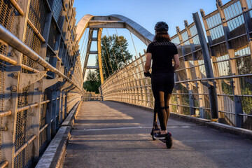 Caucasian Adult Woman riding an electric scooter on a path across a pedestrian bridge over the highway in a modern city. Taken in Fraser Heights, Surrey, Vancouver, British Columbia, Canada. © edb3_16