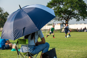 Parents sitting and watching their son playing football in a school tournament on a clear sky and sunny day. Sport, outdoor activity with lifestyle and a happy family. Soccer mom and dad concept.