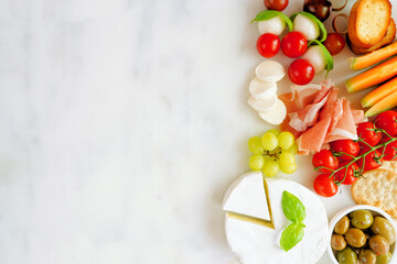 Italian theme charcuterie side border against a white marble background. Selection of cheese, meat and fruit appetizers. Overhead view with copy space.