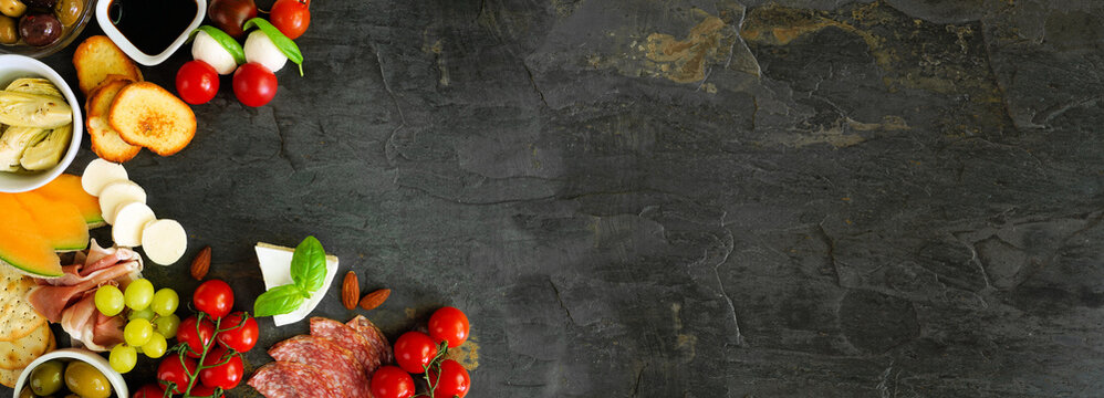 Italian theme charcuterie corner border against a dark slate banner background. Selection of cheese, meat and fruit appetizers. Above view with copy space.