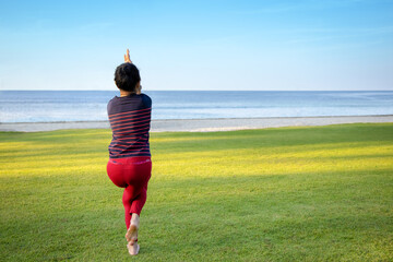Behind of senior Asian woman doing a stand yoga eagle pose (garudasana) on the grass field behind the beach and sea with clear blue sky.