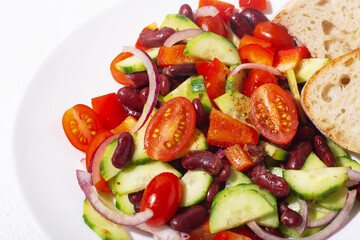 close up spanish salad with red beans and vegetables in a white plate on white background, tomatoes, cucumbers, onions, ciabatta