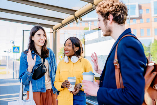 Three Multiethnic Happy Friends Meeting In Bus Stop At Street Of A Big City With An Urban Background. African And Hispanic Girl Friends Student With Curly Caucasian Man Commuting To University Togethe