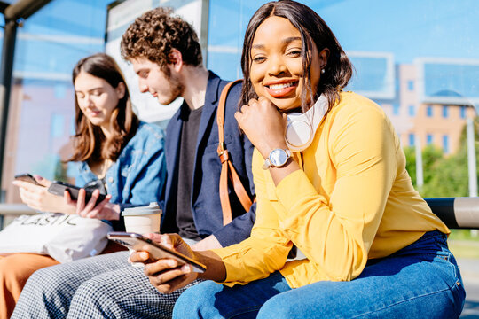 Three multiethnic friends using smart phone while sitting on the bench at bus stop and waiting for public transport at sunny bright summer day. African woman with headphones smilling at camera.