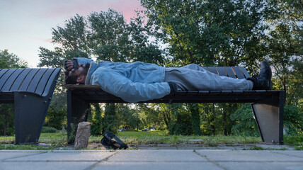 Young tired poor dirty drunk alcoholic homeless man or refugee sleeping on the wooden bench on the urban street in the city, social documentary concept