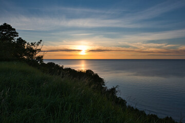 Fototapeta premium Sunset over the Baltic Sea during the white nights at the shore with a steep slope covered with grass and deciduous forest, under a sky with clouds and haze in the golden tones of the setting sun.