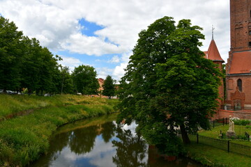 Fototapeta na wymiar A view of a vast river or lake flowing through a countryside area next to a medieval castle and a church seen next to a public park covered with many deciduous trees spotted on a cloudy summer day