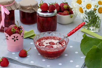 Homemade strawberry and rhubarb jam in a bowl and jars on light table in the garden.