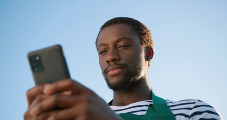 Low angle view of the optimistic multiracial farmer scientist in apron using smartphone mobile technology application while working at the field and smiling. Agriculture concept