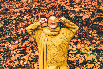 Playful young woman in bright clothes with long scarf hides eyes behind dry orange leaves near large red bush in picturesque autumn forest - Powered by Adobe