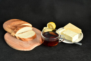 Against a black background, there is a cup of teas, a loaf of wheat bread and a plate of butter.