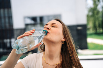 A beautiful young woman holds a plastic water bottle in her hand and drinks it outside in the heat with her eyes closed and enjoys it.