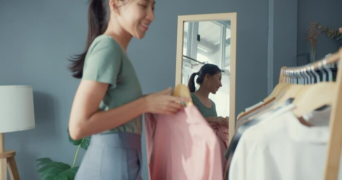 Beautiful attractive Asia lady choosing clothes on clothes rack dressing looking herself in mirror in living room at house. Girl think what to wear casual shirt. Lifestyle women relax at home concept.