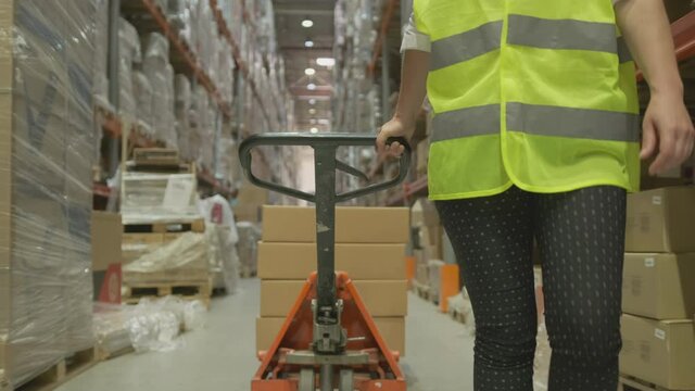 Woman storekeeper at warehouse carries hydraulic pallet truck with goods in cardboard boxes while walking between rows of shelving. Moving, shipping or acceptance of goods in logistics center.