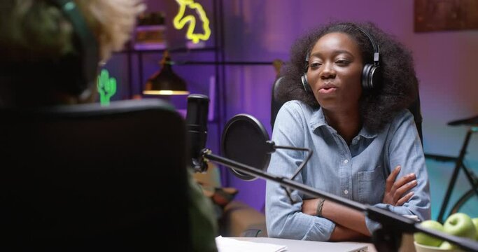Tracking shot of multiracial radio host in headphones chatting with male guest during recording of talk show in studio. Young caucasian woman listening to them talk and smiling