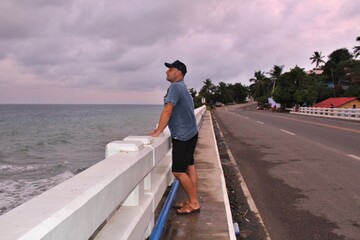 A man standing on a bridge against a gray sky and raging sea and looking into the distance